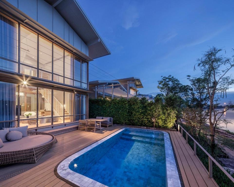 บ้านสองชั้นสมัยใหม่พร้อมหน้าต่างบานใหญ่ ระเบียงไม้ พื้นที่นั่งเล่นกลางแจ้ง และสระว่ายน้ำรูปสี่เหลี่ยมผืนผ้าในยามพลบค่ำ—เหมาะสำหรับผู้ที่มองหาที่พักอยุธยาติดแม่น้ำในสไตล์ร่วมสมัยและความสะดวกสบาย.