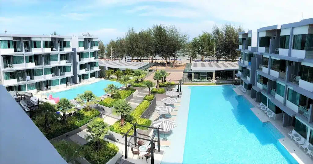 โรงแรมหัวหินสมัยใหม่ พร้อมสระว่ายน้ำสองสระ ล้อมรอบด้วยต้นไม้เขียวขจี เตียงอาบแดด ร่ม และอาคารสามชั้น—ต้นไม้และทะเลสร้างฉากหลังที่สวยงามสำหรับการเข้าพักของคุณ.