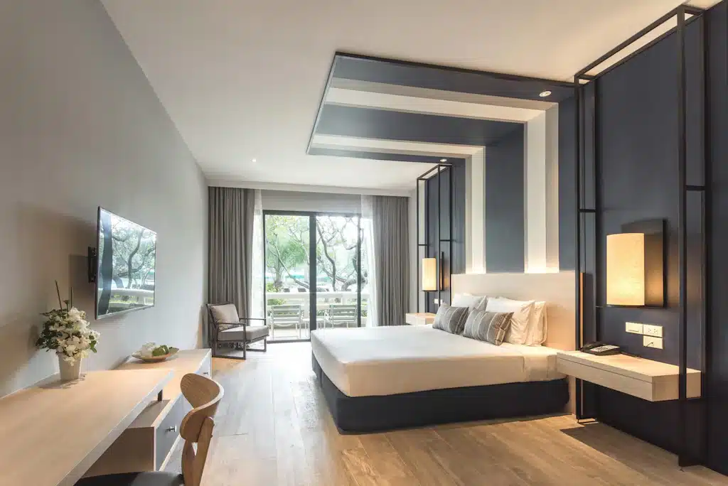 ห้องพักโรงแรมสมัยใหม่พร้อมเตียงขนาดใหญ่ ทีวีติดผนัง โต๊ะพร้อมเก้าอี้ เก้าอี้นวม และวิวจากระเบียงผ่านหน้าต่างบานใหญ่จากพื้นจรดเพดาน เหมาะสำหรับผู้ที่มองหา โรงแรมหัวหิน หรือ ที่พักหัวหินราคาถูก ในบรรยากาศที่มีสไตล์พร้อมแสงไฟร่วมสมัยและโทนสีเรียบง่าย.