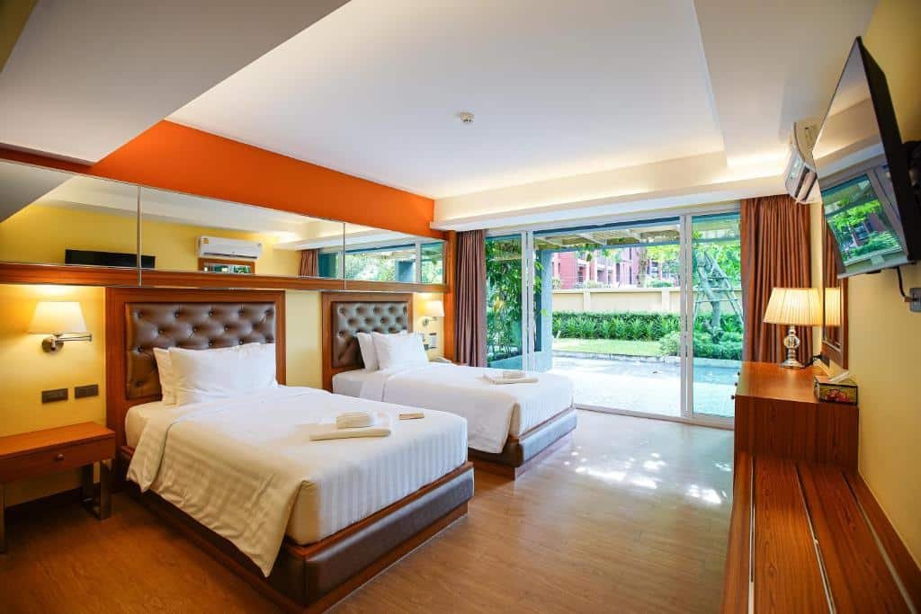 โรงแรมหัวหิน ห้องพักมีเตียงคู่สองเตียง ทีวีติดผนัง พื้นไม้ โต๊ะทำงาน และหน้าต่างสูงจรดเพดานชมวิวสวน เหมาะสำหรับผู้มองหาที่พักหัวหินราคาถูกและสะดวกสบาย.