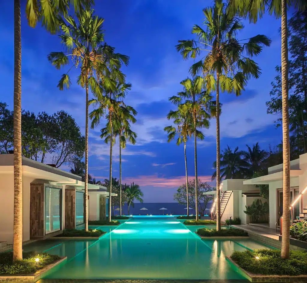 สระว่ายน้ำรูปสี่เหลี่ยมผืนผ้าที่ล้อมรอบด้วยต้นปาล์มและวิลล่าสมัยใหม่ พร้อมวิวทะเลในยามเย็น—เหมาะสำหรับผู้ที่มองหาที่พักหัวหินติดทะเลที่มีทิวทัศน์สวยงามและผ่อนคลาย.