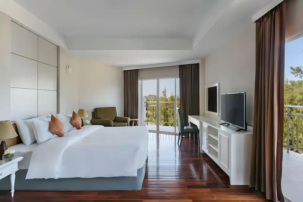 ห้องพักโรงแรมกว้างขวางพร้อมเตียงขนาดใหญ่ เก้าอี้นวม ทีวีบนโต๊ะ พื้นไม้ และระเบียงที่มองเห็นวิวทะเล เป็นตัวเลือกที่พักติดทะเลที่สมบูรณ์แบบในหัวหิน—ผ้าม่านสีน้ำตาลล้อมกรอบประตูบานเลื่อนกระจกเพื่อให้แสงธรรมชาติเข้ามาอย่างเต็มที่.