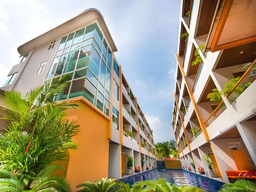 อาคารอพาร์ตเมนต์สมัยใหม่พร้อมระเบียงมองเห็นสระว่ายน้ำ ล้อมรอบด้วยพืชเขตร้อนใต้ท้องฟ้าที่มีเมฆบางส่วน—เป็นตัวเลือกที่เหมาะสำหรับผู้ที่ต้องการที่พักหัวหินใกล้ทะเล หรือโรงแรมหัวหิน สำหรับการพักผ่อนที่ผ่อนคลาย.