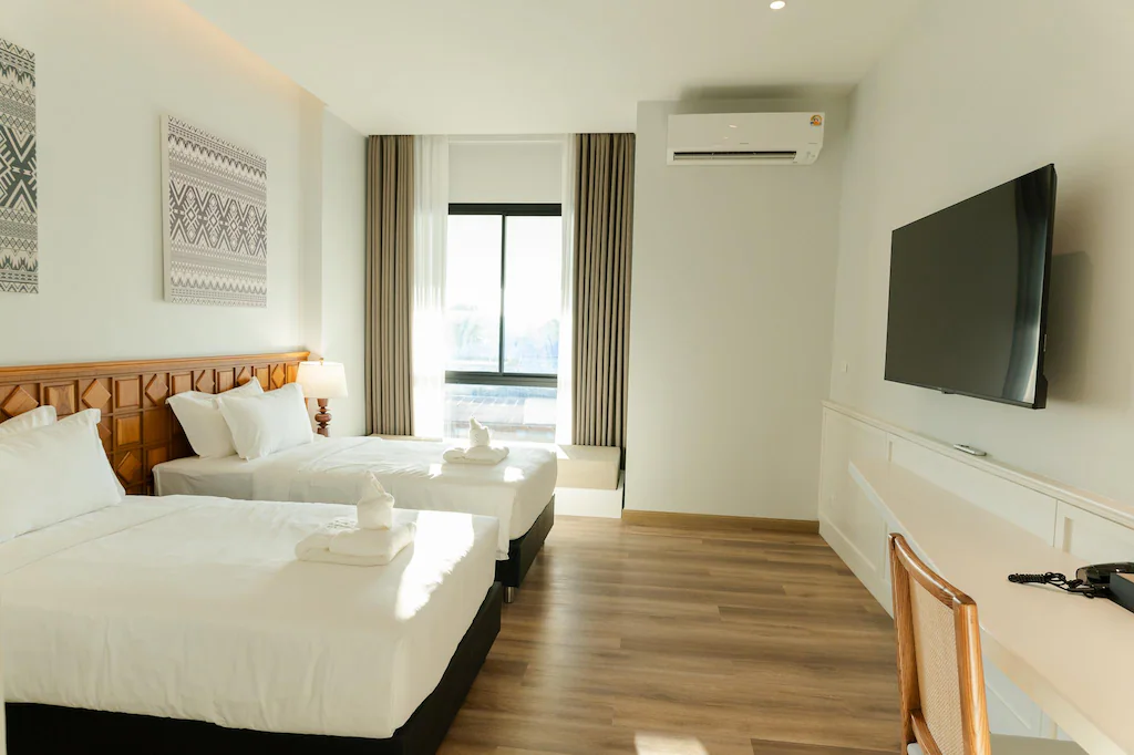 ห้องพักโรงแรมพร้อมเตียงคู่สองเตียง โทรทัศน์ติดผนัง โต๊ะพร้อมเก้าอี้ เครื่องปรับอากาศ และหน้าต่างพร้อมผ้าม่านที่เปิดรับแสงธรรมชาติ—เหมาะสำหรับผู้ที่มองหา ที่พักในเมืองน่าน ที่สะดวกสบายและครบครัน.