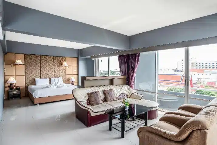 ห้องพักโรงแรมกว้างขวางพร้อมเตียงขนาดใหญ่ โซฟา 2 ตัว โต๊ะกาแฟกระจก และหน้าต่างบานใหญ่จากพื้นจรดเพดานที่มองเห็นตึกในเมือง—เหมาะสำหรับการเข้าพักของคุณที่ โรงแรมอยุธยา หรือใครก็ตามที่กำลังมองหา ที่พักอยุธยา ที่มีสไตล์.