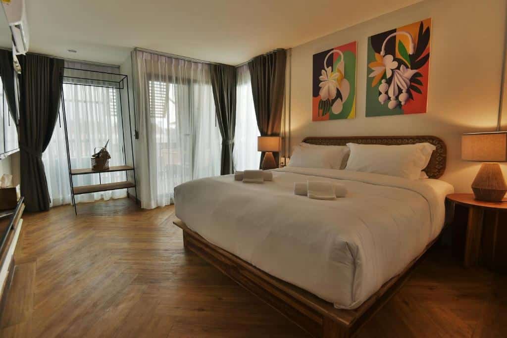 เตียงที่จัดไว้อย่างเรียบร้อยพร้อมผ้าปูที่นอนสีขาวในห้องโรงแรมอยุธยาที่ทันสมัย มีพื้นไม้ โคมไฟข้างเตียงสองข้าง งานศิลปะดอกไม้สีสันสดใส และหน้าต่างบานใหญ่พร้อมผ้าม่าน.
