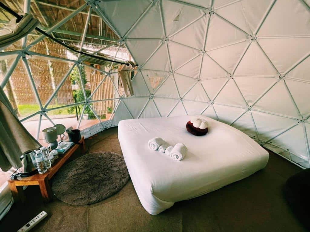 เตียงที่มีผ้าเช็ดตัวม้วนวางอยู่ด้านในห้องพักทรงโดมจีโอเดสิกสีขาว ที่พักบ่อเกลือ พร้อมโต๊ะเล็ก ขวดน้ำ แก้ว และหน้าต่างที่มองเห็นวิวต้นไม้เขียวชอุ่ม.
