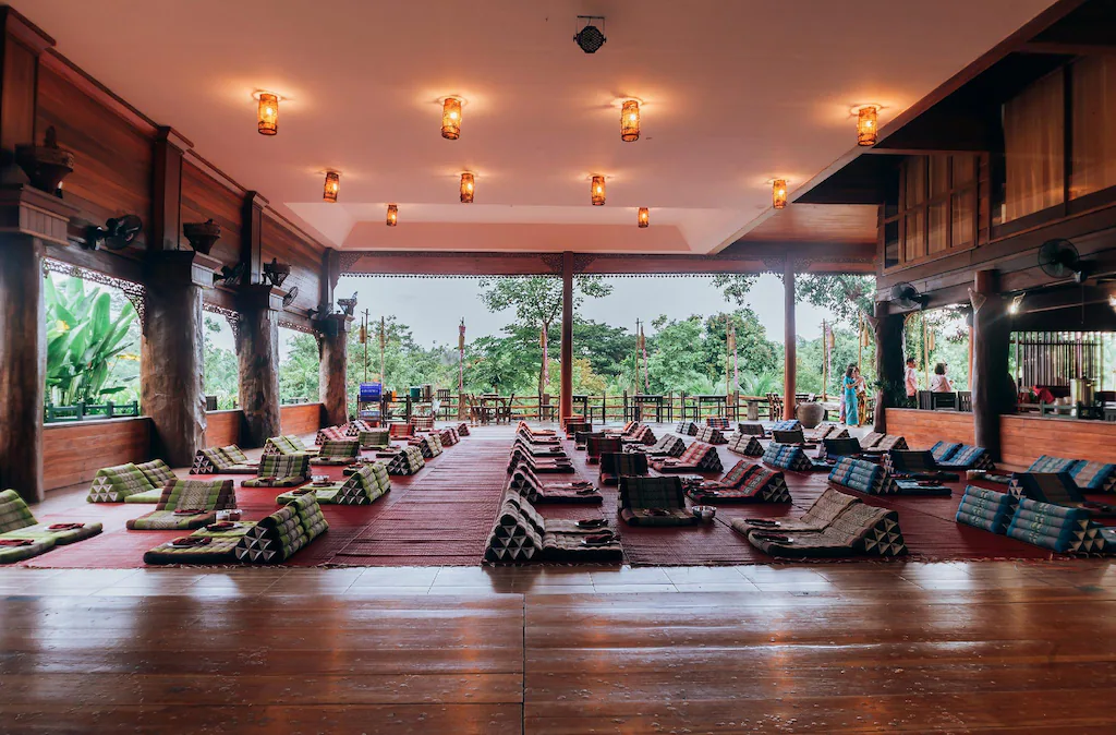 ห้องโล่งกว้างขวางพร้อมพื้นไม้ เสื่อต่ำ และเบาะรองนั่งเรียงเป็นแถว มองเห็นพื้นที่สีเขียวผ่านผนังเปิดโล่ง เหมาะสำหรับการพักผ่อนที่ ที่พักน่าน หรือ ที่พักบ่อเกลือ พร้อมไฟเพดานที่สร้างบรรยากาศอบอุ่น.