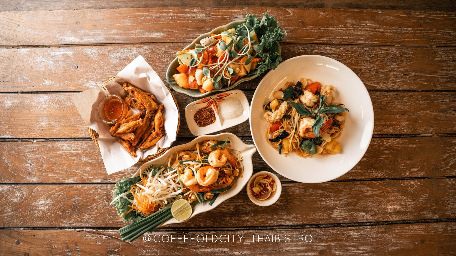 มุมมองจากด้านบนของอาหารไทยหลากหลายชนิด รวมถึงกุ้ง, บะหมี่, ปีกไก่, และผักสดบนโต๊ะไม้สไตล์บ้านๆ—เหมาะสำหรับผู้ที่วางแผนจะไปเที่ยวอยุธยาและเพลิดเพลินกับรสชาติท้องถิ่นที่แท้จริง.