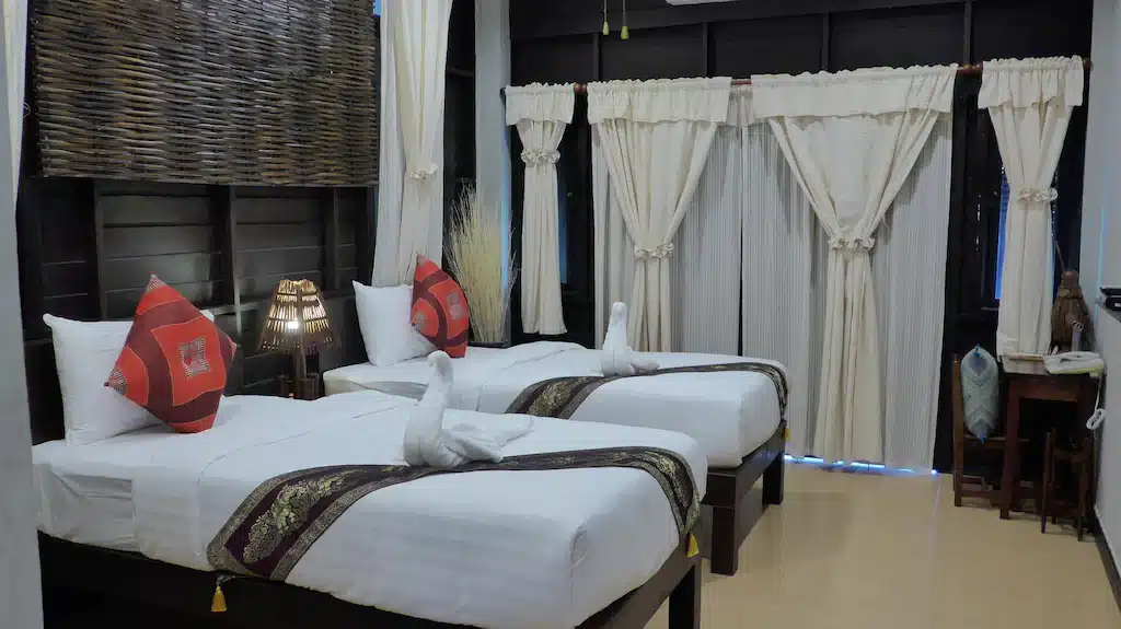 เตียงเดี่ยวสองเตียงพร้อมผ้าปูที่นอนสีขาวและหมอนตกแต่งในห้องที่มีผนังไม้สีเข้ม ผ้าม่านสีครีม และโต๊ะทำงานขนาดเล็ก—เหมาะสำหรับการพักผ่อนหลังจากสำรวจ โรงแรมอยุธยา หรือเพลิดเพลินกับการเข้าพักที่ ที่พักอยุธยาริมน้ำ.