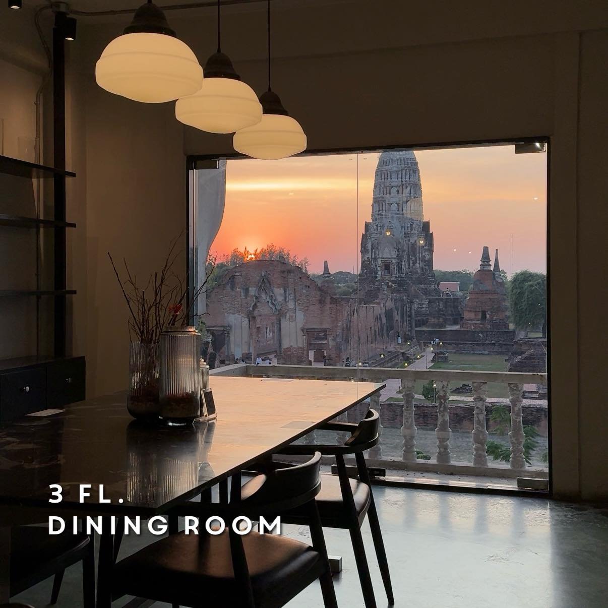 ห้องรับประทานอาหารบนชั้นสามพร้อมหน้าต่างบานใหญ่ที่มองเห็นซากปรักหักพังของวัดโบราณยามพระอาทิตย์ตก—จุดที่สวยงามสำหรับผู้มาเยือนอยุธยาที่เที่ยว. โคมไฟแขวนสไตล์โมเดิร์นแขวนอยู่เหนือโต๊ะพร้อมเก้าอี้ เพิ่มสไตล์ให้กับการเดินทางของคุณ.