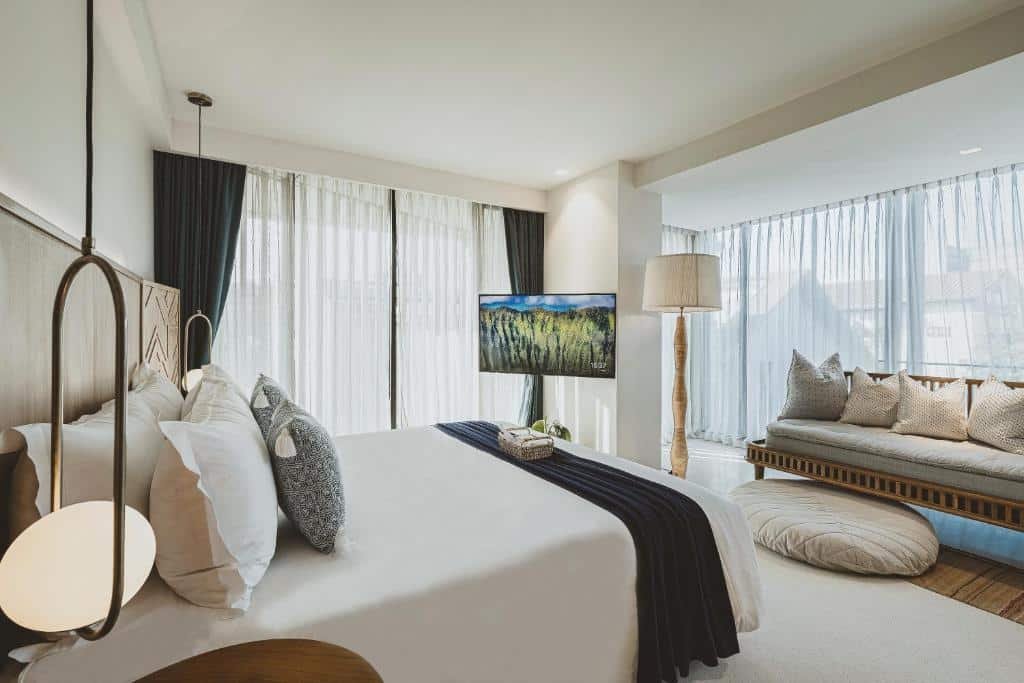 ห้องพักโรงแรมสมัยใหม่พร้อมเตียงขนาดใหญ่ หน้าต่างบานใหญ่จากพื้นจรดเพดาน โทรทัศน์ โซฟาพร้อมหมอน และโคมไฟตั้งพื้นในบรรยากาศสว่างไสวและมินิมอล—เหมาะสำหรับการพักผ่อนหลังจากเที่ยวเชียงใหม่หรือสำรวจสถานที่ท่องเที่ยวเชียงใหม่ยอดนิยม.