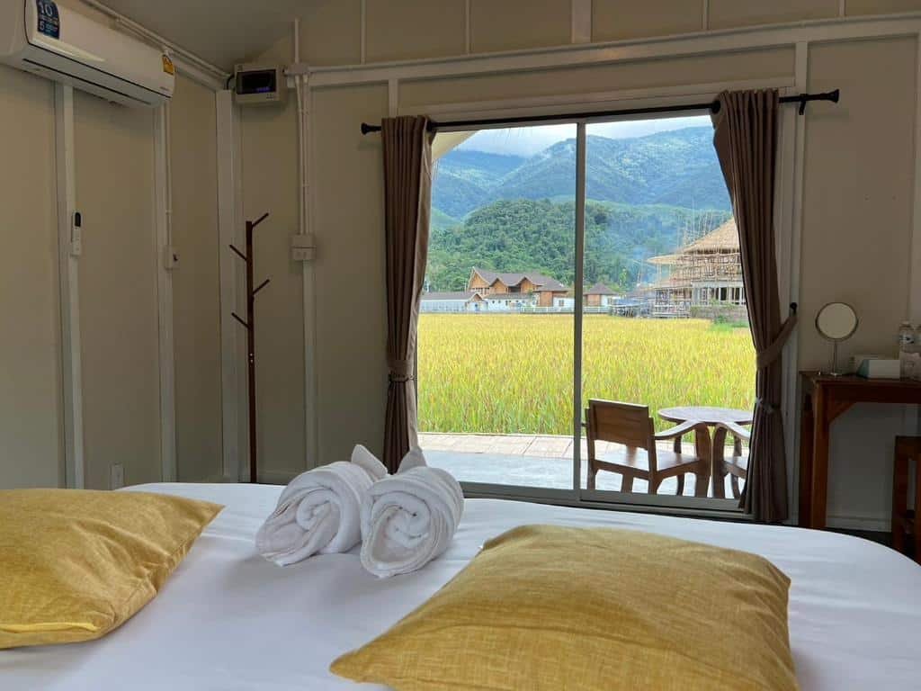 ห้องพักโรงแรมที่มีเตียงปูผ้าเรียบร้อย หมอนสีเหลืองสองใบ และศิลปะจากผ้าขนหนู มองออกไปผ่านประตูกระจกสู่ลานระเบียง ทุ่งหญ้าสีเขียว และภูเขา—เหมาะสำหรับการพักผ่อนที่ ที่พักน่าน ใจกลางเมือง.