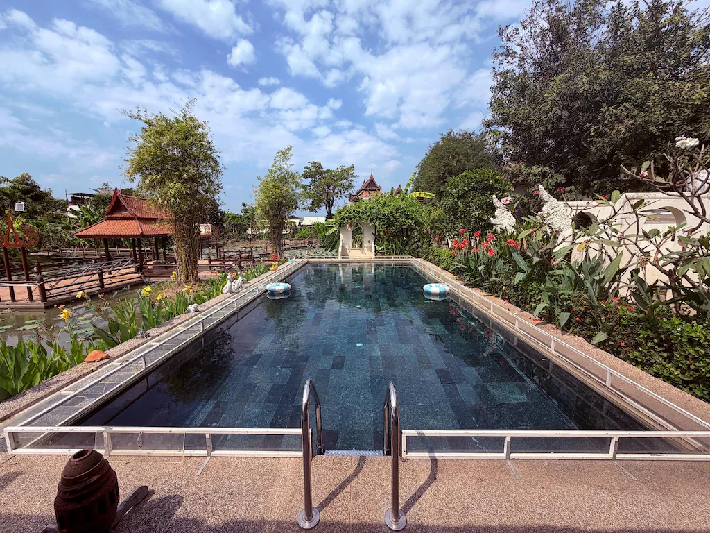 สระว่ายน้ำกลางแจ้งรูปสี่เหลี่ยมผืนผ้าพร้อมราวจับโลหะ ล้อมรอบด้วยพืชและต้นไม้เขตร้อน ตั้งอยู่ใต้ท้องฟ้าสีครามที่มีเมฆกระจายอยู่เป็นระยะ—เหมาะอย่างยิ่งสำหรับการพักผ่อนที่ ที่พักอยุธยาริมน้ำ หรือ โรงแรมอยุธยา.