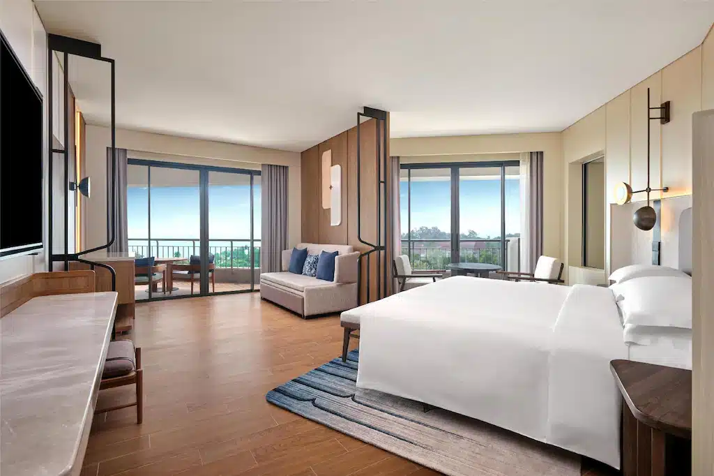 ห้องพักโรงแรมกว้างขวางที่โรงแรมหัวหิน พร้อมเตียงขนาดใหญ่ โซฟา โต๊ะทำงาน และประตูกระจกสองบานที่นำไปสู่ระเบียงพร้อมที่นั่งกลางแจ้งและวิวสวนสีเขียว เหมาะสำหรับใครที่กำลังมองหาที่พักหัวหินราคาประหยัดและสะดวกสบาย.