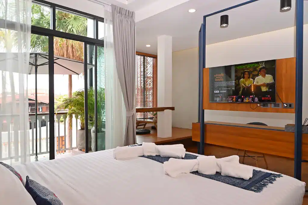 ห้องพักโรงแรมสมัยใหม่พร้อมเตียงขนาดใหญ่ ผ้าเช็ดตัวพับเรียบร้อย โต๊ะทำงาน โทรทัศน์ติดผนัง และระเบียงพร้อมที่นั่งกลางแจ้งและต้นไม้เขตร้อน—เหมาะสำหรับการพักผ่อนที่ ที่พักน่านในเมือง.