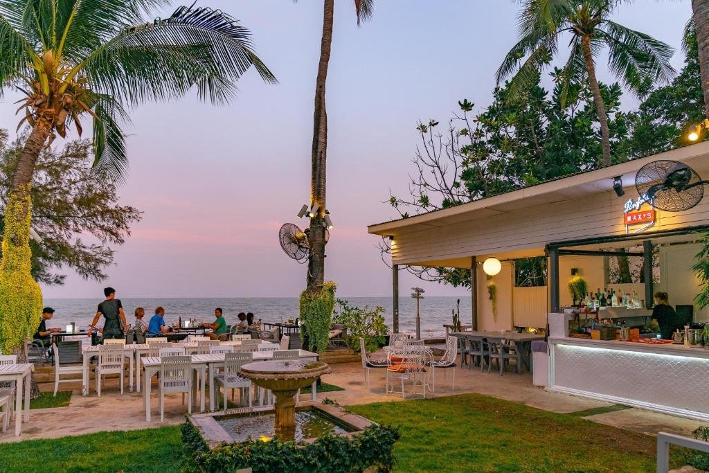 ร้านอาหารริมทะเลกลางแจ้งที่มีโต๊ะและเก้าอี้สีขาว น้ำพุขนาดเล็ก ต้นปาล์ม และผู้คนกำลังรับประทานอาหารยามพระอาทิตย์ตกดิน บริเวณบาร์ที่มีแสงไฟส่องสว่างมองเห็นวิวทะเล เหมาะอย่างยิ่งสำหรับแขกที่กำลังมองหาที่พักริมทะเลหัวหินพร้อมวิวที่สวยงามตระการตา.