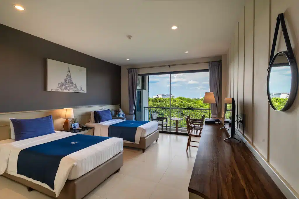 ห้องพักโรงแรมสมัยใหม่พร้อมเตียงสองเตียง ผ้าปูที่นอนสีน้ำเงินและขาว โต๊ะไม้ขนาดใหญ่ กระจกกลม และระเบียงที่มองเห็นทิวทัศน์สีเขียวภายใต้ท้องฟ้าสีคราม—เหมาะสำหรับการเข้าพักของคุณที่ โรงแรมอยุธยา.