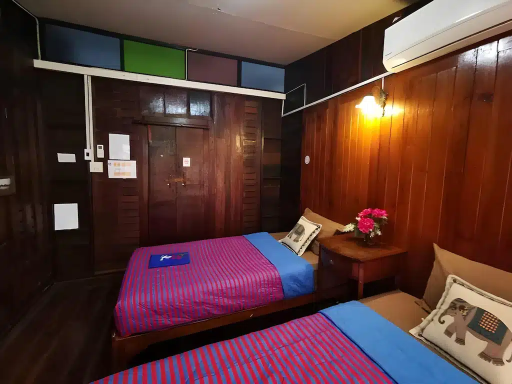 เตียงเดี่ยวสองเตียงพร้อมผ้าปูที่นอนลายทางในห้องที่ตกแต่งด้วยไม้ที่ โรงแรมอยุธยา พร้อมโต๊ะข้างเตียงที่มีดอกไม้ หมอนประดับ โคมไฟติดผนัง และเครื่องปรับอากาศเพื่อความสะดวกสบายของคุณ.