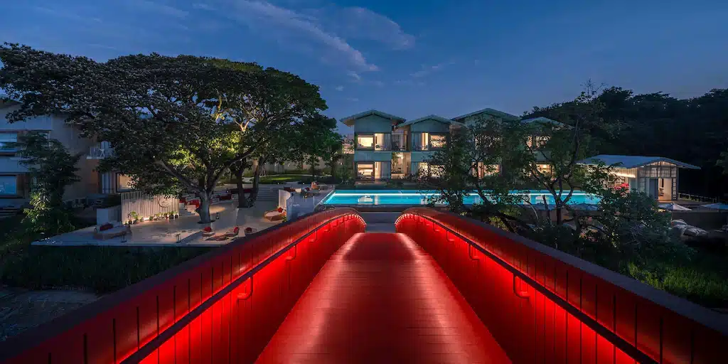 สะพานสีแดงส่องสว่างทอดยาวไปยังบ้านสมัยใหม่ที่มีหน้าต่างบานใหญ่และสระว่ายน้ำ ล้อมรอบด้วยต้นไม้ในยามพลบค่ำ สร้างความรู้สึกเสน่ห์ของโรงแรมอยุธยาที่ซ่อนตัวอยู่ในธรรมชาติที่เงียบสงบ.