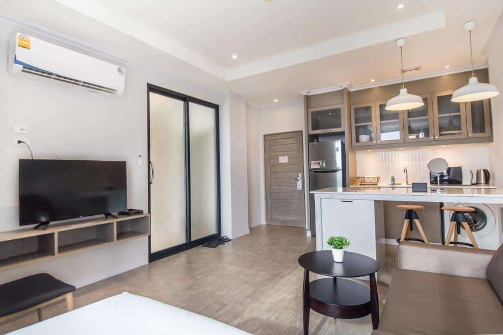 อพาร์ตเมนต์สตูดิโอสมัยใหม่พร้อมครัวขนาดเล็ก เก้าอี้บาร์ ทีวีติดผนัง เครื่องปรับอากาศ และโต๊ะรับประทานอาหารขนาดเล็กพร้อมต้นไม้ เหมาะสำหรับนักเดินทางที่กำลังมองหาที่พักราคาถูกในหัวหินในพื้นที่เงียบสงบด้วยโทนสีเรียบและการตกแต่งแบบมินิมอล.