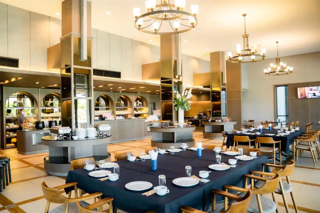 พื้นที่รับประทานอาหารของร้านอาหารสมัยใหม่ที่โรงแรมอยุธยา มีโต๊ะจัดเตรียมสำหรับมื้ออาหาร เก้าอี้ไม้ ผ้าปูโต๊ะสีดำ และเคาน์เตอร์บุฟเฟ่ต์อยู่ด้านหลังภายใต้โคมไฟระย้าที่สว่างไสว.