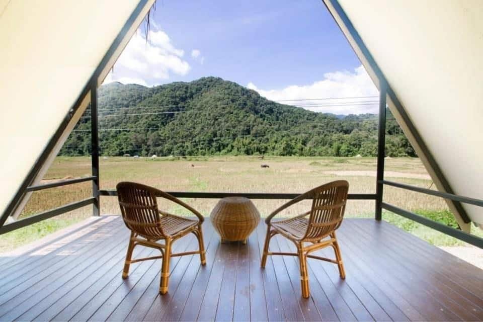 เก้าอี้หวายสองตัวและโต๊ะกลมขนาดเล็กบนดาดฟ้าไม้ในโครงสร้างรูปตัว A ที่ที่พักปัว มองเห็นทุ่งนาและเนินเขาสีเขียวภายใต้ท้องฟ้าสีคราม.