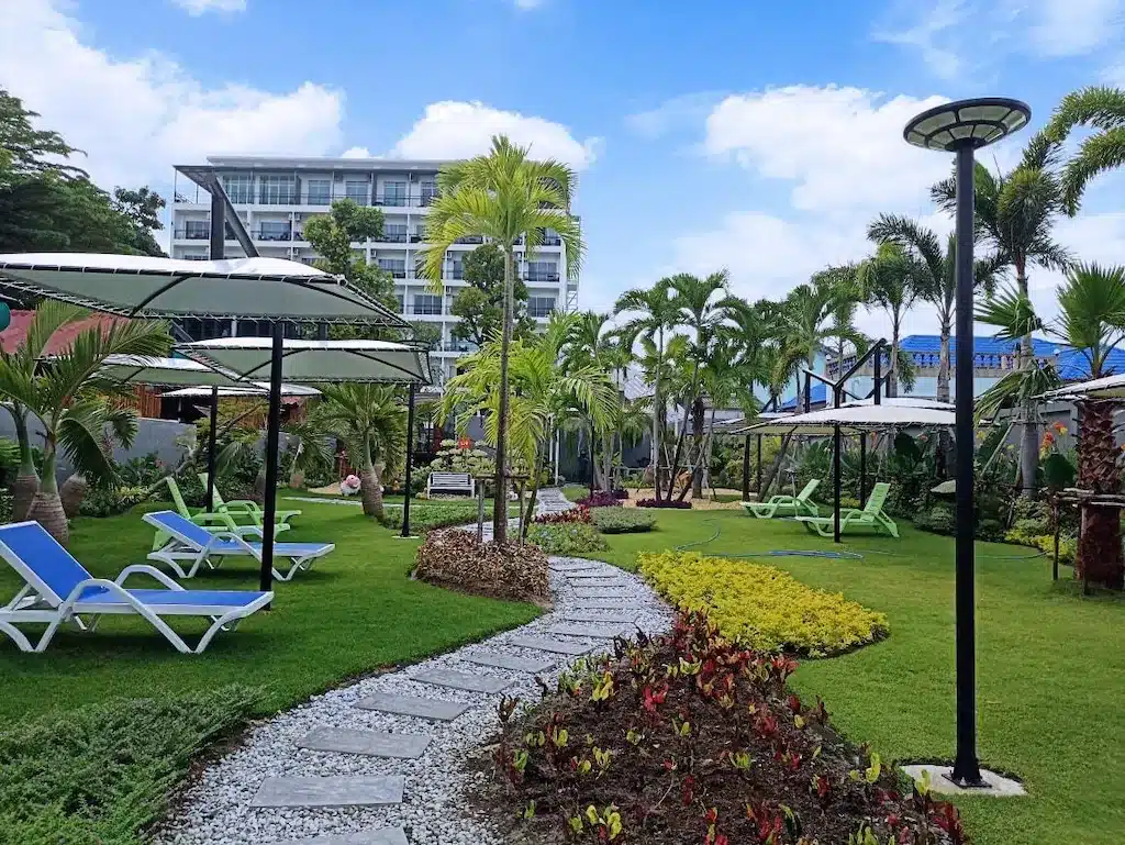 สวนที่จัดแต่งอย่างสวยงามพร้อมเก้าอี้พักผ่อน ร่ม ต้นปาล์ม ทางเดินหิน และโรงแรมหัวหินสมัยใหม่หลายชั้นเป็นฉากหลังภายใต้ท้องฟ้าสีคราม.