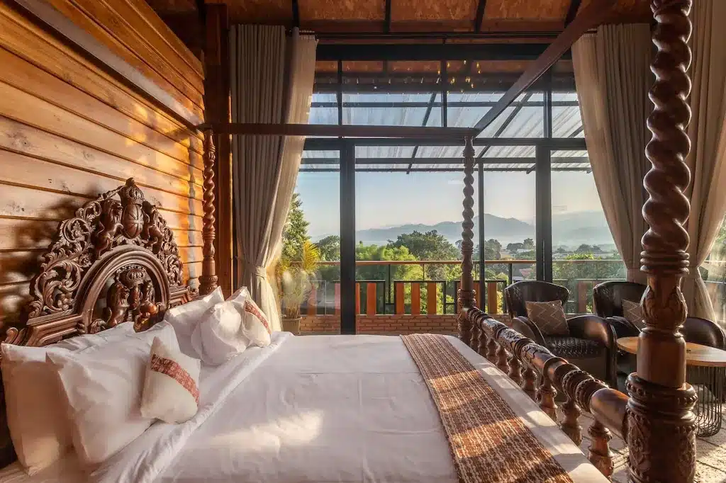 ห้องนอนที่มีเตียงไม้แกะสลัก ผ้าปูที่นอนสีขาว หน้าต่างบานใหญ่ ม่าน และเก้าอี้หวาย—ที่พักแห่งนี้ในน่านมีระเบียงที่มองเห็นต้นไม้และภูเขาไกลๆ เพื่อการพักผ่อนอย่างสงบ.