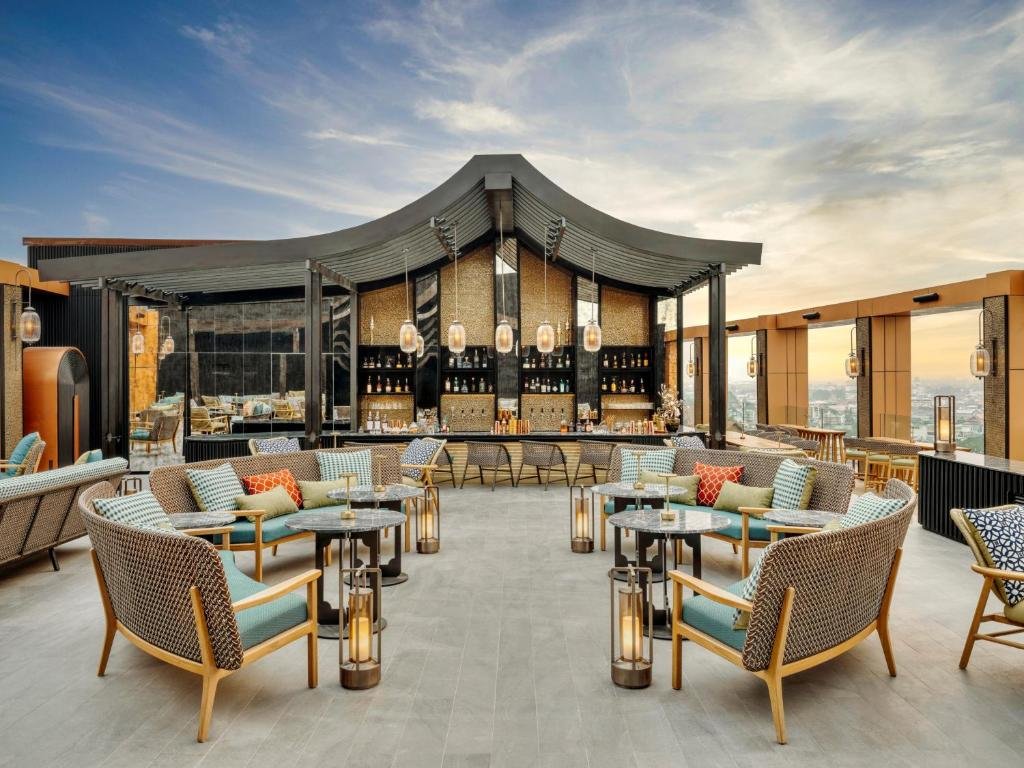 บาร์บนดาดฟ้าที่มีที่นั่งโค้ง โต๊ะสไตล์โมเดิร์น โคมไฟแขวน และพื้นที่บาร์กลาง พร้อมวิวทิวทัศน์ของเมืองยามพระอาทิตย์ตก—เหมาะสำหรับผู้ที่มองหาสถานที่ท่องเที่ยวเชียงใหม่ในสไตล์ที่หรูหรา.