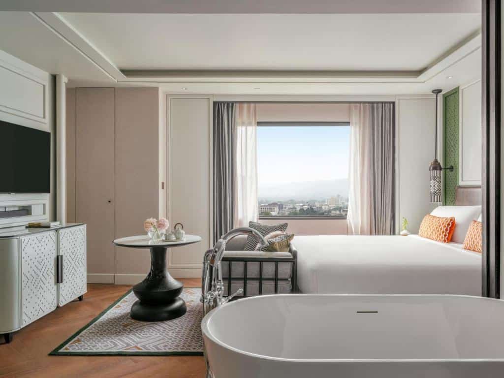 ห้องพักโรงแรมสมัยใหม่พร้อมเตียงขนาดใหญ่ ทีวีติดผนัง โต๊ะกลมพร้อมเก้าอี้ และอ่างอาบน้ำแบบลอยตัว มีหน้าต่างที่มองเห็นวิวเมืองและภูเขา เหมาะสำหรับการพักผ่อนหลังจากเที่ยวเชียงใหม่ทั้งวัน.