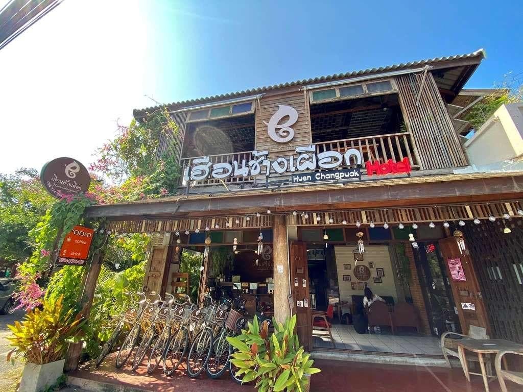 ภาพถนนหน้าทางเข้าโรงแรมฮุยช้างเผือก ที่พักน่ารักในเมืองน่าน พร้อมจักรยานจอดอยู่ พืชพรรณเขียวชอุ่ม และผนังไม้ภายนอกภายใต้ท้องฟ้าแจ่มใส.