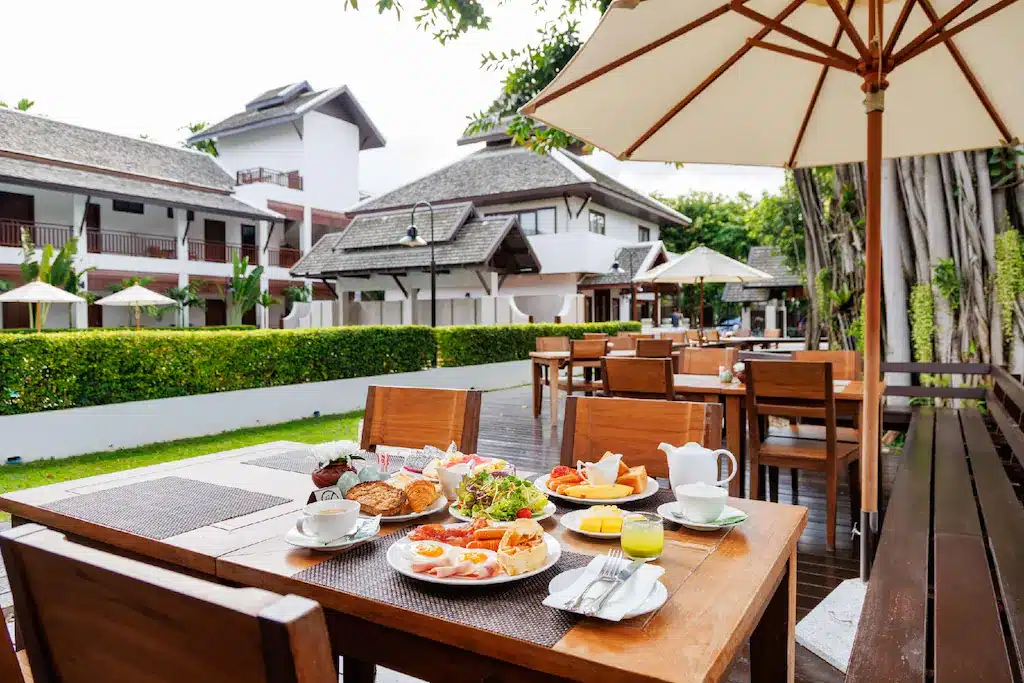 พื้นที่รับประทานอาหารกลางแจ้งที่รีสอร์ทในเชียงใหม่ หนึ่งในสถานที่ท่องเที่ยวเชียงใหม่ยอดนิยม พร้อมโต๊ะไม้สำหรับอาหารเช้า—ขนมอบ ไข่ ผลไม้ และเครื่องดื่ม—ทั้งหมดล้อมรอบด้วยพืชพรรณเขียวชอุ่มและอาคารสีขาวอันมีเสน่ห์.