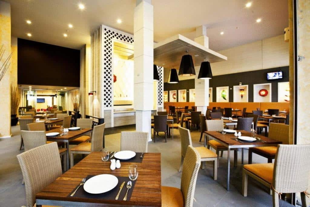 ร้านอาหารสมัยใหม่ภายในตกแต่งด้วยโต๊ะและเก้าอี้ว่างเปล่า แสงไฟอบอุ่น และของตกแต่งร่วมสมัย—เหมาะสำหรับแขกจาก โรงแรมหัวหิน ที่ต้องการประสบการณ์การรับประทานอาหารอย่างมีสไตล์ งานศิลปะบนผนังและโทรทัศน์ช่วยเสริมบรรยากาศที่น่าดึงดูดใจ.