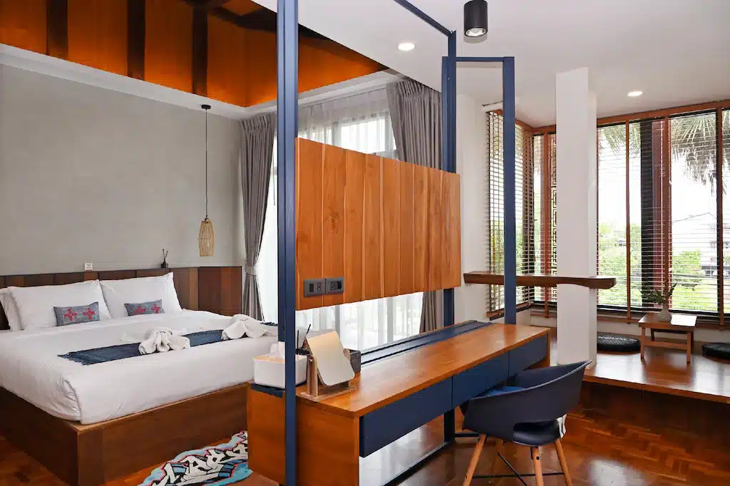 ห้องพักโรงแรมสมัยใหม่พร้อมเตียงขนาดใหญ่ โต๊ะทำงาน และเก้าอี้ มีฉากกั้นไม้ หน้าต่างบานใหญ่พร้อมม่านบังแสง และพื้นที่นั่งเล่นขนาดเล็ก—เหมาะสำหรับนักเดินทางที่กำลังมองหาที่พักในเมืองน่านหรือที่พักสไตล์ทันสมัยในน่าน.