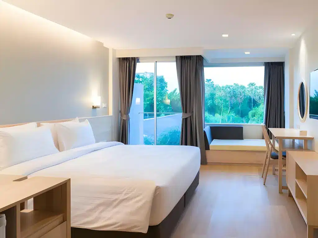 ห้องพักโรงแรมสมัยใหม่ที่ โรงแรมหัวหิน พร้อมเตียงขนาดใหญ่ โต๊ะทำงาน โทรทัศน์ติดผนัง และหน้าต่างบานใหญ่จากพื้นจรดเพดานที่มองเห็นวิวสีเขียว; ตกแต่งสไตล์มินิมอลด้วยเฟอร์นิเจอร์ไม้สีอ่อนและโทนสีกลางเพื่อการพักผ่อนที่ผ่อนคลาย.