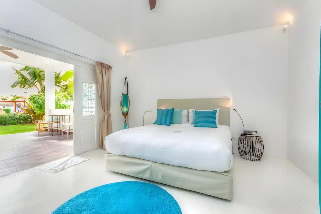 ห้องนอนสีขาวทันสมัยพร้อมเตียงขนาดใหญ่ หมอนอิงสีเขียวอมฟ้า พรมกลมสีเขียวอมฟ้า และประตูบานเลื่อนที่เปิดออกสู่ลานกลางแจ้งที่มีต้นไม้สีเขียว—เหมาะสำหรับผู้ที่มองหา โรงแรมหัวหิน หรือ ที่พักหัวหิน ที่มีสไตล์.