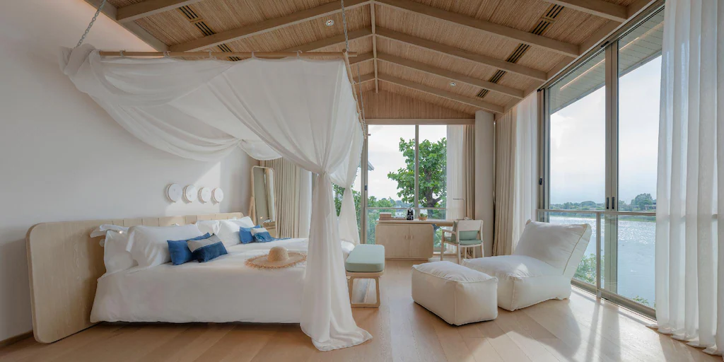 ห้องนอนสมัยใหม่พร้อมเตียงหลังคาผ้าคลุม ผ้าปูที่นอนสีขาว หมอนอิงสีน้ำเงิน โซนพักผ่อน และเพดานไม้ หน้าต่างบานใหญ่เปิดรับวิวของน้ำและต้นไม้เขียวขจี—เหมาะสำหรับผู้ที่ต้องการที่พักริมน้ำในบรรยากาศเงียบสงบในอยุธยา.