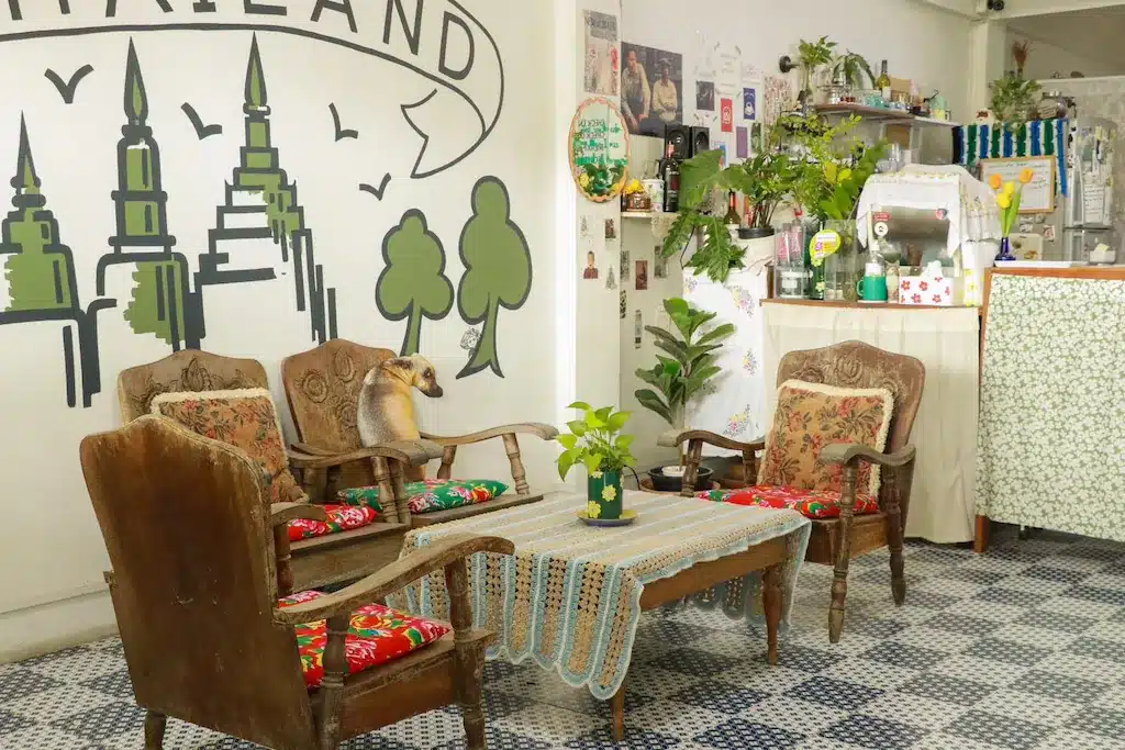 คาเฟ่บรรยากาศอบอุ่นพร้อมเก้าอี้ไม้สไตล์วินเทจ โต๊ะปูผ้าลูกไม้ ต้นไม้กระถาง ศิลปะบนผนังเป็นภาพวัดไทย และเคาน์เตอร์อยู่ด้านหลัง—เหมาะสำหรับการพักผ่อนหลังจากเที่ยวชม ที่พักอยุธยา หรือ โรงแรมอยุธยา ในบริเวณใกล้เคียง.