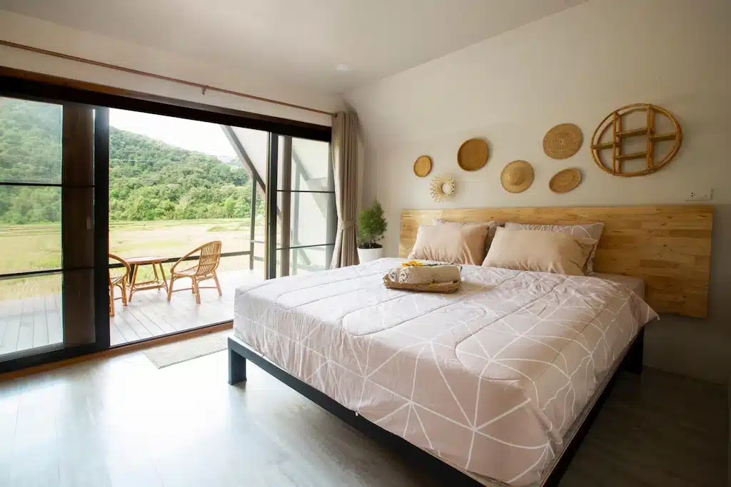 ห้องนอนสมัยใหม่พร้อมเตียงขนาดใหญ่ ตกแต่งผนัง และประตูบานเลื่อนกระจกที่เปิดออกสู่ระเบียงซึ่งมีเก้าอี้สองตัวและโต๊ะ มองเห็นทิวทัศน์สีเขียว เหมาะสำหรับการพักผ่อนที่ ที่พักน่านในเมือง.