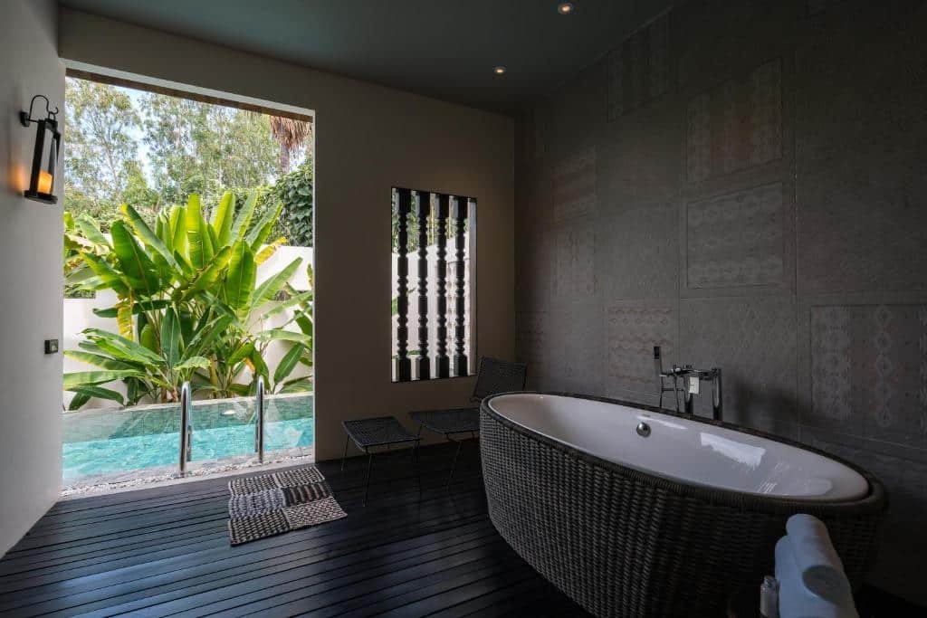 ห้องน้ำสมัยใหม่ที่ โรงแรมอยุธยา พร้อมอ่างอาบน้ำแบบลอยตัว เก้าอี้สองตัว และวิวสระว่ายน้ำกลางแจ้งและต้นไม้เขียวชอุ่มผ่านผนังเปิดโล่ง.