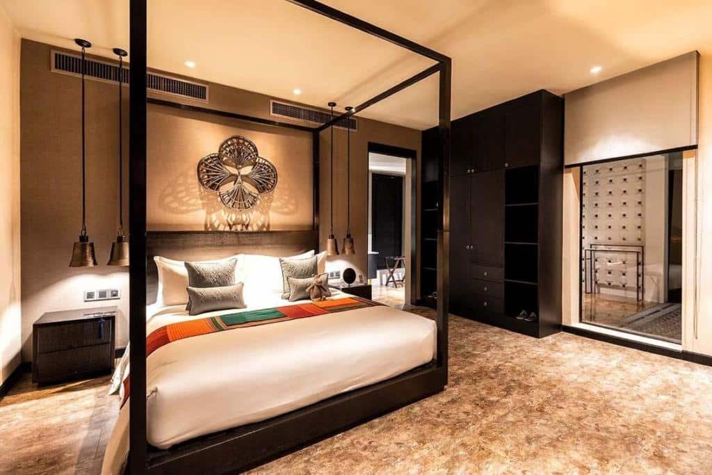 ห้องนอนสมัยใหม่พร้อมเตียงสี่เสา ศิลปะตกแต่งผนัง โคมไฟแขวน และตู้เสื้อผ้าบานกระจกขนาดใหญ่ คล้ายกับห้องที่มีสไตล์ที่ โรงแรมอยุธยา มีแสงไฟอบอุ่นและพื้นลายเพิ่มเสน่ห์.