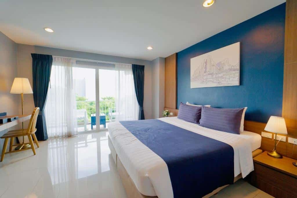 ห้องพักโรงแรมสมัยใหม่ที่ โรงแรมอยุธยา พร้อมเตียงขนาดใหญ่ ผนังสีน้ำเงินเป็นจุดเด่น โต๊ะทำงาน โคมไฟ และระเบียงพร้อมประตูกระจกที่เปิดรับแสงธรรมชาติ—เหมาะสำหรับการเข้าพักของคุณในอยุธยา.