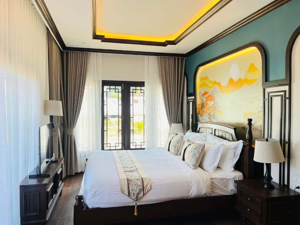 เตียงที่จัดไว้อย่างเรียบร้อยพร้อมผ้าปูที่นอนสีขาวในห้องนอนที่สว่างสดใส มีภาพจิตรกรรมฝาผนังเหนือหัวเตียง โคมไฟสองดวง ผ้าม่าน และทีวีบนขาตั้งใกล้หน้าต่างสูง—เหมาะสำหรับการพักผ่อนหลังจากสำรวจสถานที่ท่องเที่ยวในอยุธยา.