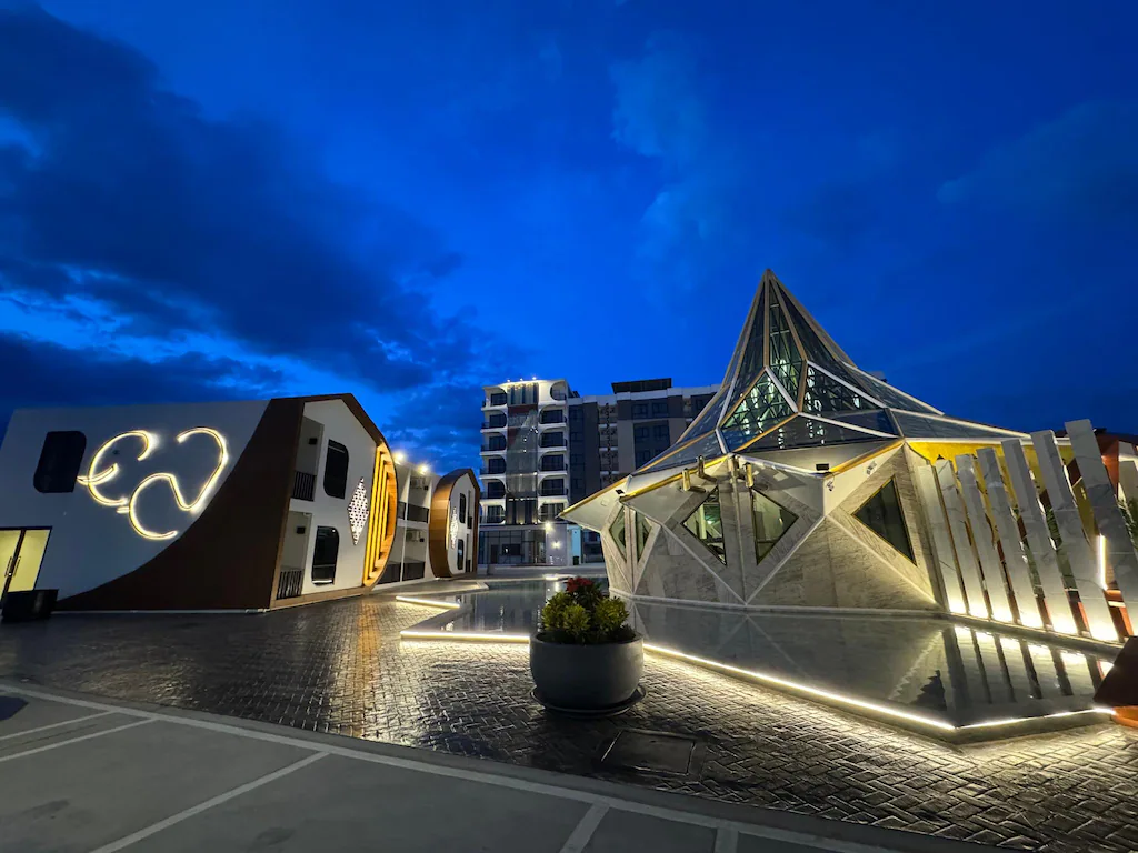 อาคารสมัยใหม่ที่มีการออกแบบเชิงเรขาคณิตและเส้นขอบที่ส่องสว่างตั้งอยู่ใต้ท้องฟ้ายามค่ำคืนสีน้ำเงินเข้ม โดยมีต้นไม้ในกระถางอยู่ด้านหน้า—เป็นแรงบันดาลใจที่สมบูรณ์แบบสำหรับ โรงแรมอยุธยา ที่มีสไตล์ใจกลางเมือง.