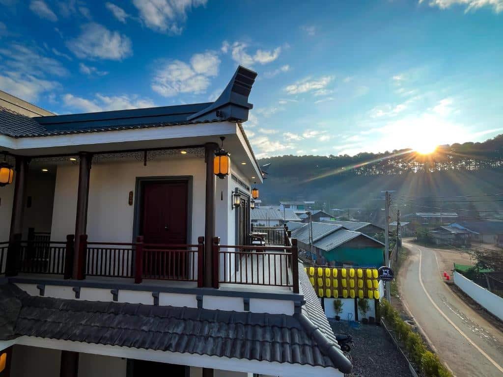 อาคารสองชั้นที่มีสถาปัตยกรรมแบบดั้งเดิมตั้งตระหง่านมองเห็นถนนในหมู่บ้านยามพระอาทิตย์ขึ้น โดยมีต้นไม้และเนินเขาเป็นกรอบภาพ—ถ่ายทอดเสน่ห์อันเงียบสงบของสถานที่ท่องเที่ยวอยุธยาภายใต้ท้องฟ้าที่มีเมฆบางส่วน.