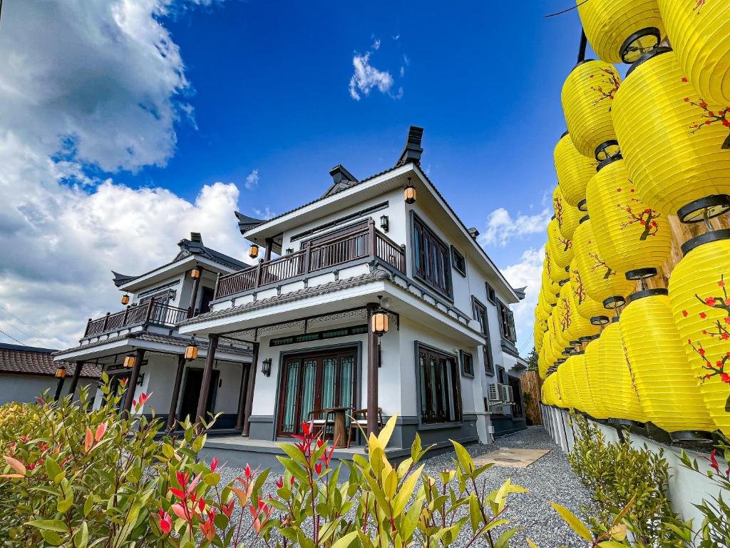 อาคารสีขาวสองชั้นพร้อมขอบสีเข้มตั้งอยู่ข้างกำแพงประดับโคมไฟสีเหลืองใต้ท้องฟ้าสีฟ้าที่มีเมฆกระจายอยู่เป็นระยะ มอบเสน่ห์คลาสสิกท่ามกลางสถานที่ท่องเที่ยวอยุธยา พุ่มไม้ตั้งอยู่ด้านหน้า.