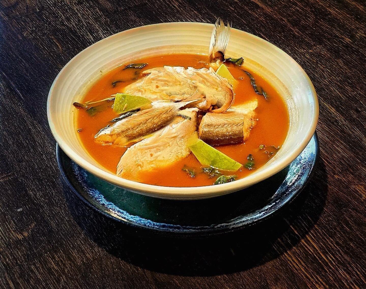 ชามซุปปลาพร้อมชิ้นปลา มะนาวฝาน และสมุนไพรในน้ำซุปสีส้ม เสิร์ฟบนโต๊ะไม้สีเข้ม—เหมาะสำหรับลิ้มรสหลังจากวันที่สำรวจสถานที่ท่องเที่ยวอยุธยา.