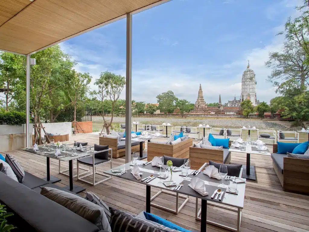 ลานระเบียงร้านอาหารกลางแจ้งพร้อมโต๊ะที่จัดเรียงอย่างเรียบร้อยและเก้าอี้ที่มีเบาะรองนั่ง มองเห็นแม่น้ำและโครงสร้างวัดโบราณภายใต้ท้องฟ้าที่มีเมฆบางส่วน—เหมาะสำหรับแขกของที่พักอยุธยาริมน้ำหรือโรงแรมอยุธยาที่ต้องการความผ่อนคลาย.