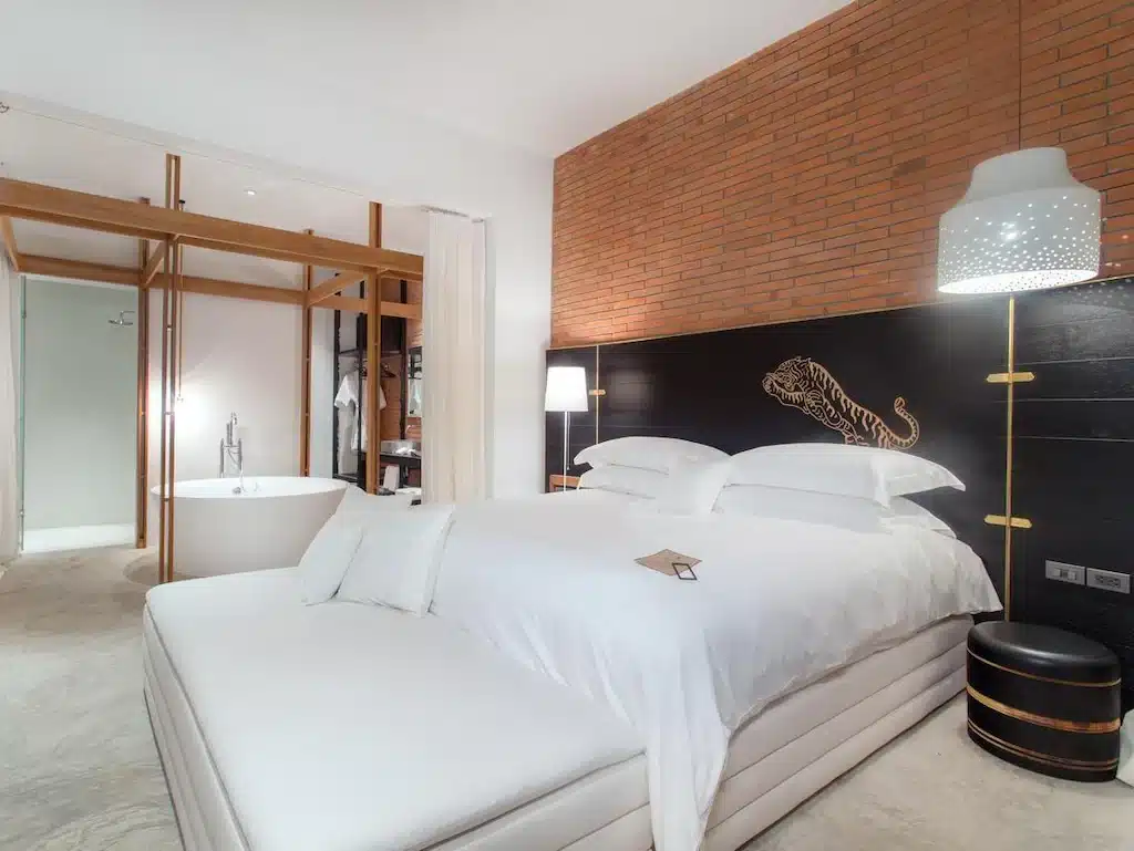 ห้องพักโรงแรมสมัยใหม่พร้อมเตียงขนาดใหญ่ เครื่องนอนสีขาว ผนังตกแต่งด้วยอิฐ ห้องน้ำพร้อมอ่างอาบน้ำแบบลอยตัว และไฟส่องสว่างสไตล์ร่วมสมัย—เหมาะสำหรับผู้ที่มองหาที่พักในอยุธยาที่ทั้งมีสไตล์และสะดวกสบาย.